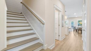 A staircase with wooden steps and a handrail, leading to a hallway with a door and a view of a dining area with a table and chairs. - Large Home~Short Walk to Beach+Saltwater Pool l Sleeps 24 (Panama City Beach)