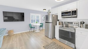 Une cuisine moderne avec des meubles blancs, des appareils en acier inoxydable, un coin repas avec une table ronde et des chaises, et un téléviseur à écran plat fixé au mur.