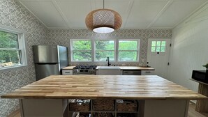 Een keuken met een centraal eiland, een fornuis, een koelkast en een spoelbak. Drie ramen zorgen voor natuurlijk licht, en er hangt een lamp boven het eiland.