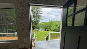 Uitzicht vanuit een kamer met behang met een patroon en een houten vloer, op een terras met een hekwerk en een gazon met bomen.
