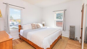A bedroom with a wooden bed, a bedside table, a radiator, and a window with curtains. - Modern Loft in Historic Birdtown - Unit 8 (Lakewood)