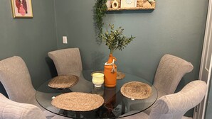 Zona de menjador amb taula de vidre envoltada per quatre cadires amb coixins, un suport per plats trenat, un vas amb una planta i una estant decorativa amb una imatge i un vas petit.