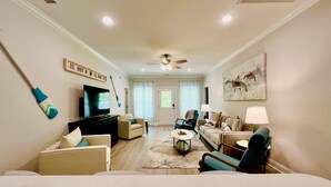 A spacious living room with a flat-screen TV, comfortable seating, a ceiling fan, and a large window with curtains. - Private Pool, Pet-Friendly, New 3BR Home in Cherry Grove (North Myrtle Beach)