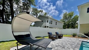 A patio with a swing chair, two wicker chairs, a pool, and a white fence. - Private Pool, Pet-Friendly, New 3BR Home in Cherry Grove (North Myrtle Beach)