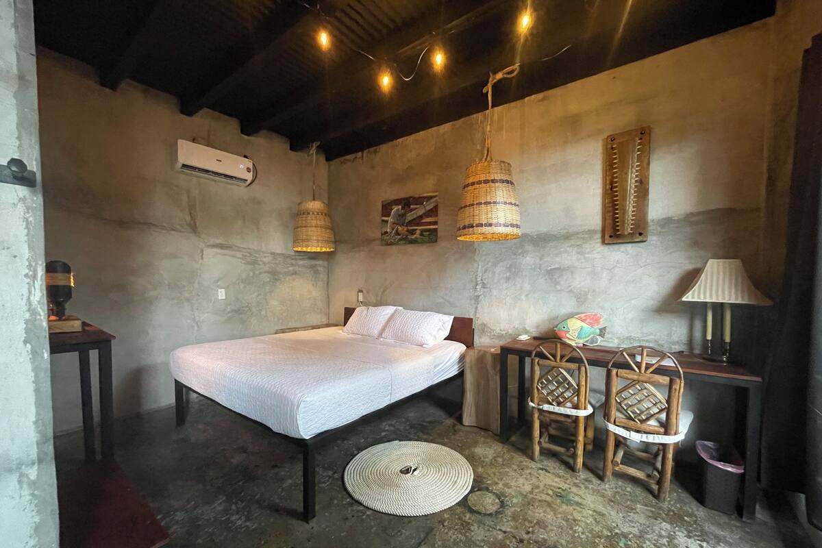 A rustic bedroom with a single bed, a small table, two chairs, a lamp, and a hanging light fixture.