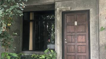 A building with a large wooden door, a window with vertical blinds, and a small porch area with greenery.