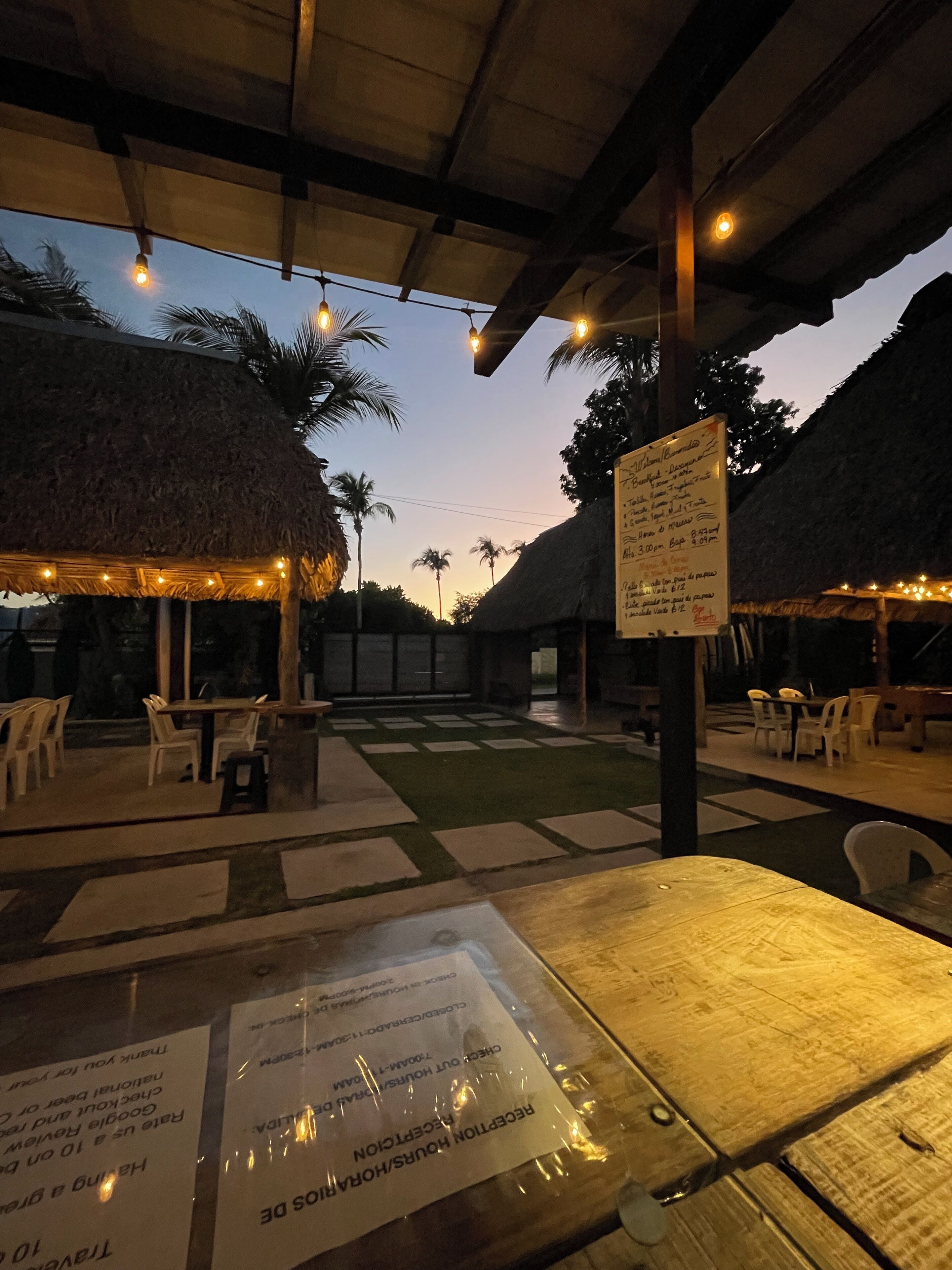 An outdoor dining area with tables and chairs, a thatched-roof structure, and a menu sign.