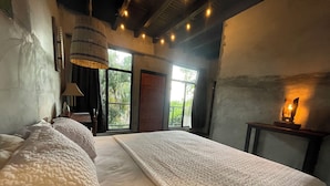 A bedroom with a large bed, a nightstand, a lamp, and a view of greenery outside.