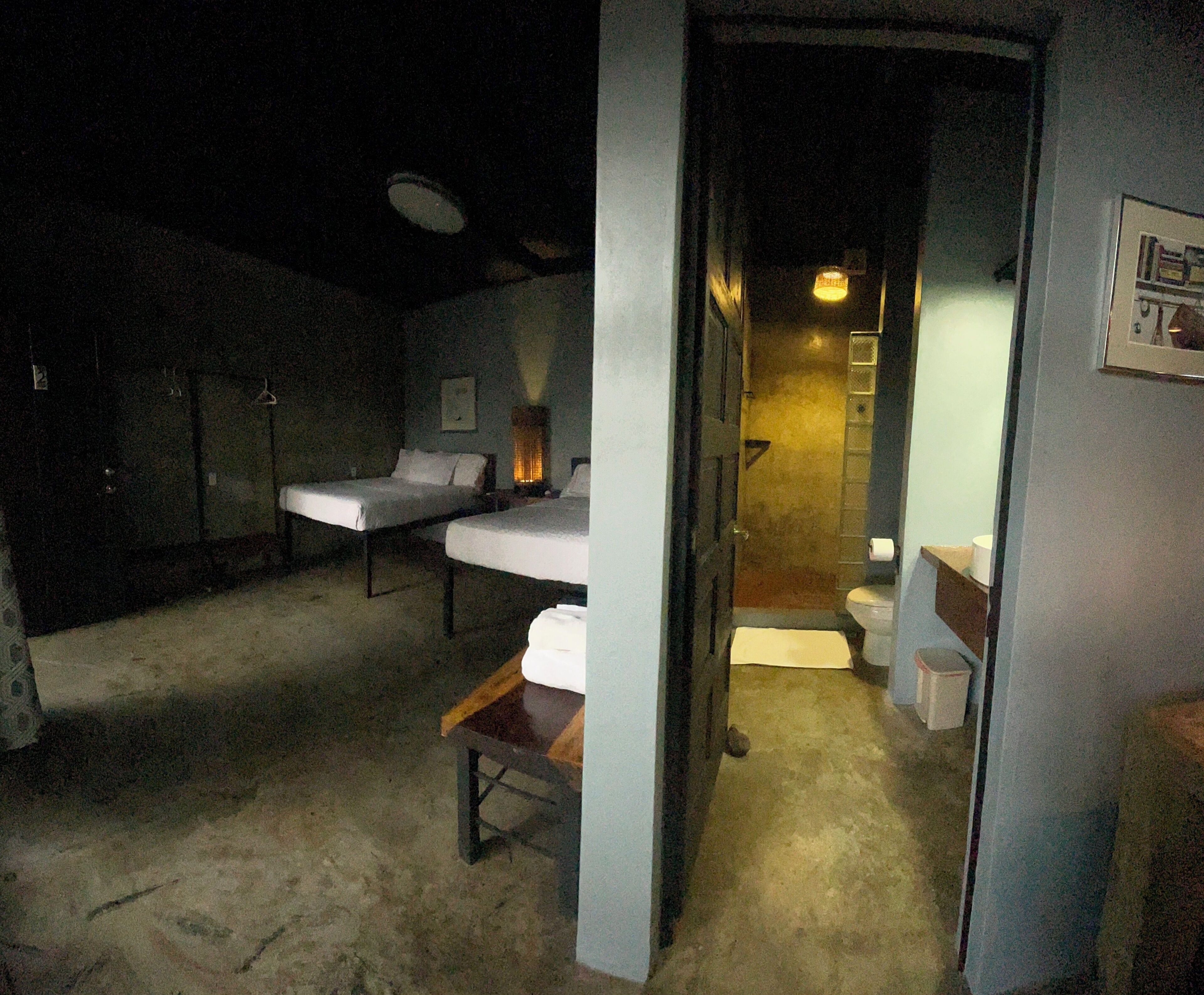 A dimly lit room with two single beds, a small table, and a door leading to a bathroom.
