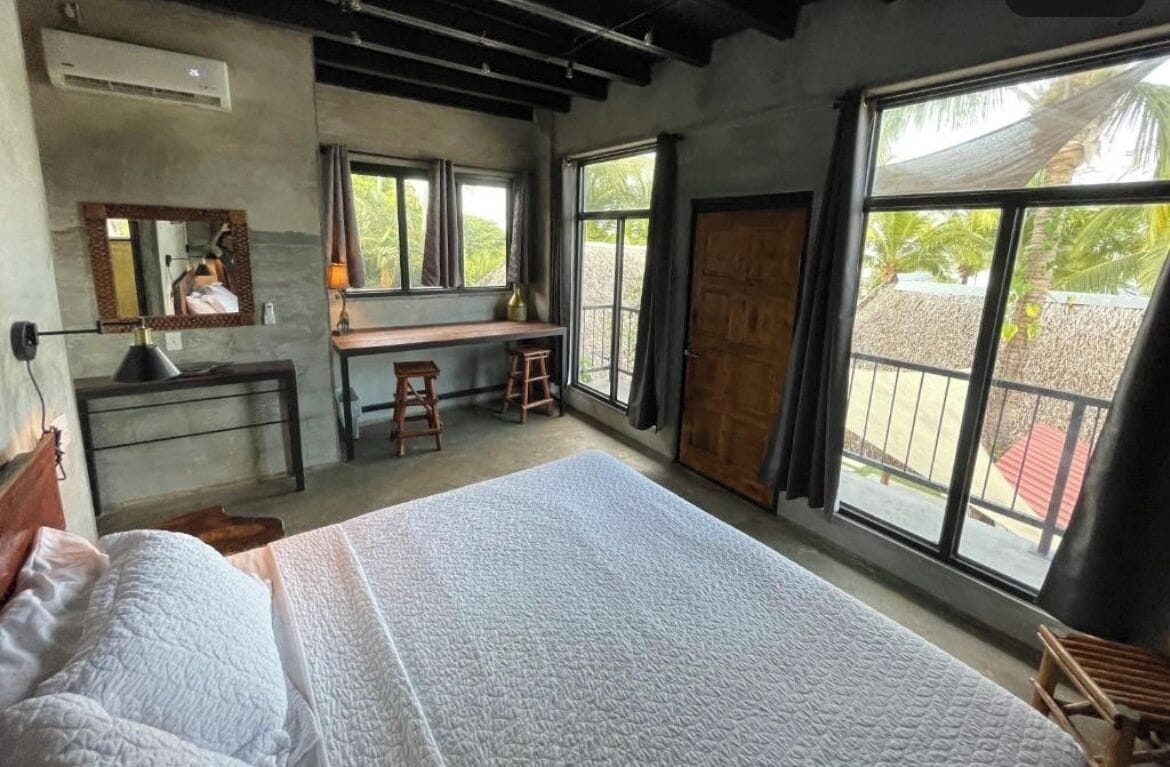 Een slaapkamer met een groot bed, een bureau met twee krukken, een spiegel en grote ramen met uitzicht op palmbomen.