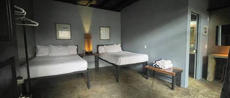 Una habitación con dos camas, un perchero, una mesita de noche con lámpara y un pequeño banco con toallas.