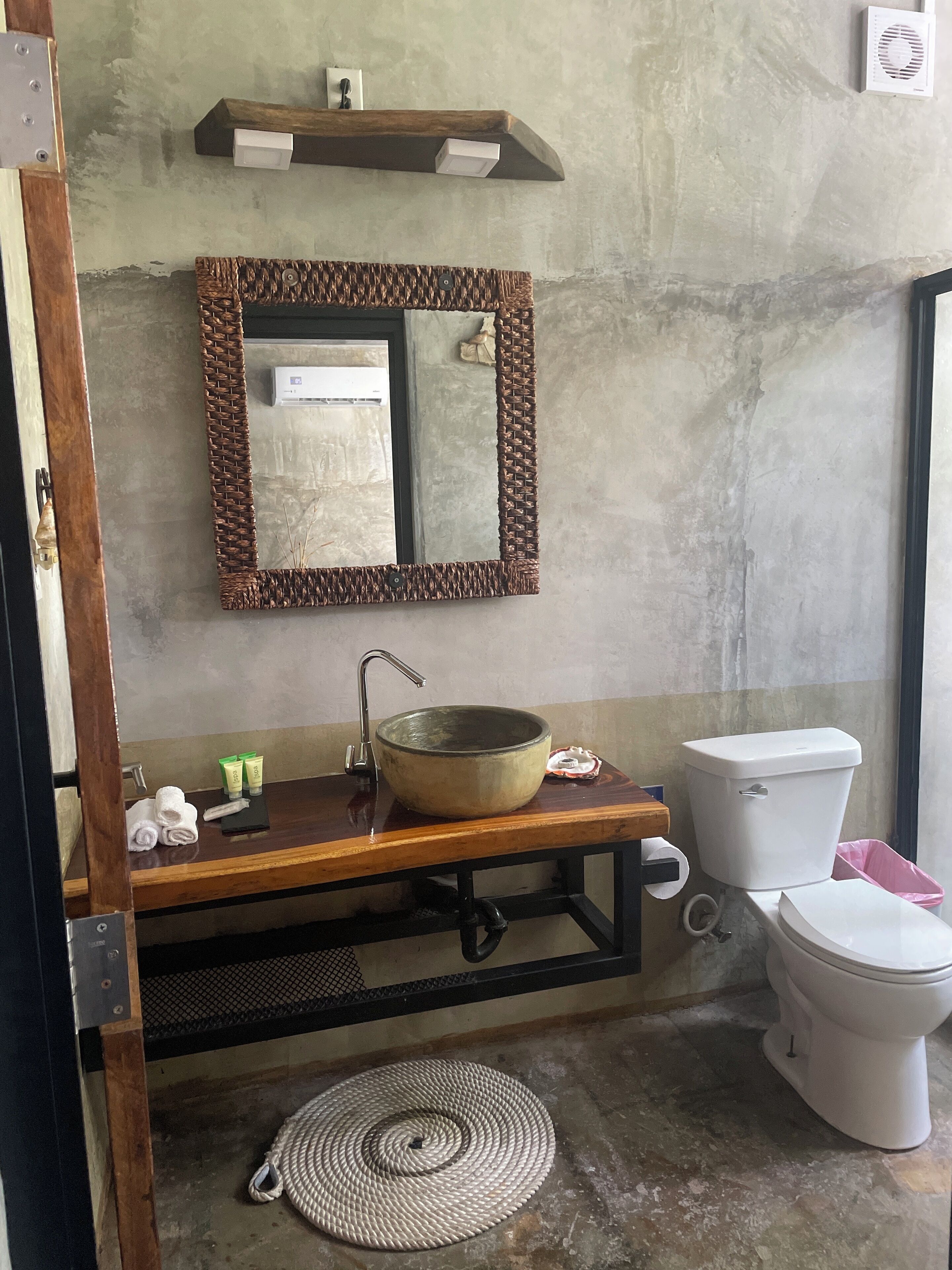Ein Badezimmer mit einem Holzkosmetikschrank, einem großen Spiegel mit Geweberahmen, einer weißen Toilette und einer gewebten Matte auf dem Boden.