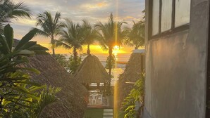 A staircase leading to a terrace with a view of the sunset over the ocean, surrounded by palm trees and thatched structures.