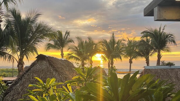 A beach scene with palm trees and thatched huts, a sunset in the background, and a concrete structure to the right.