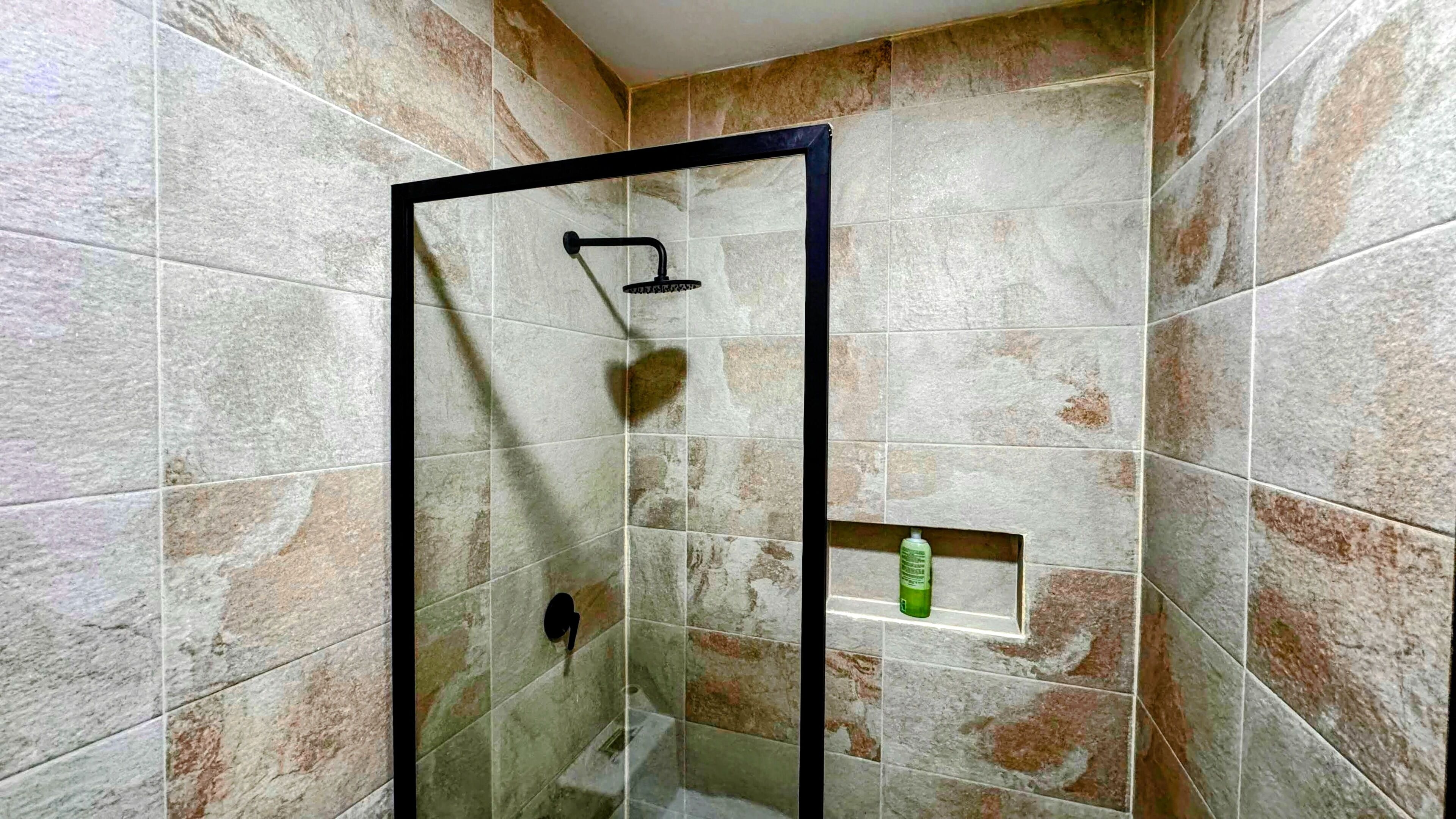 A glass-enclosed shower with a black frame, beige and brown tiles, and a green bottle on a shelf.