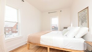 Een slaapkamer met een houten bed, wit beddengoed, een raam met uitzicht op gebouwen en een kunstwerk in een lijst aan de muur.