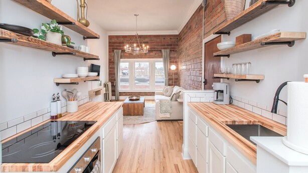 Een moderne keuken met houten aanrecht, een ingebouwde kookplaat en een metselwerk muur. Er zijn planken met keukengerei en een gezellige zithoek met een bank en een bijzettafeltje.
