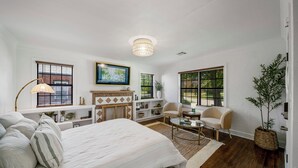 A spacious bedroom with a large bed, a desk, two chairs, a fireplace, and a large window with blinds.