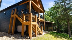 Una cabaña de madera de dos pisos con balcón y escaleras, rodeada de árboles.
