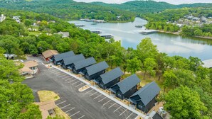 Un hotel con varios edificios de techos oscuros, un estacionamiento y una vista panorámica de un lago con barcos y una ladera boscosa al fondo.