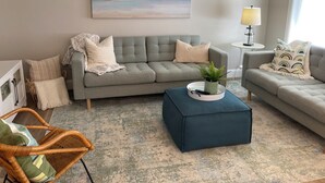A living room with a grey sofa, a blue ottoman, a wicker chair, a small table with a lamp, and a painting of a seascape. - Cozy all season retreat, center of town! (Garden City)