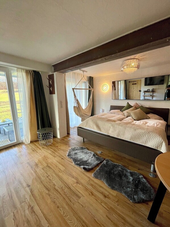 Une chambre moderne avec un grand lit, un fauteuil suspendu, une petite table ronde et une porte-fenêtre coulissante donnant sur un espace extérieur.