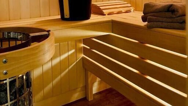 Una sauna con bancos de madera y un asiento en banco, un cubo negro y una toalla sobre el banco.