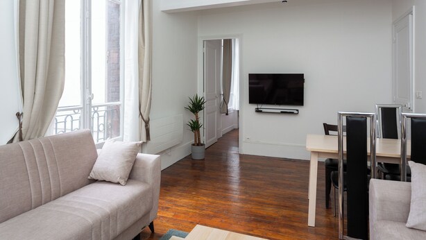A modern living room with a sofa, a dining table with chairs, a TV mounted on the wall, and a potted plant. - Cosy Marx Dormoy apartment at the foot of the metro  (Paris)