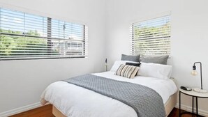 A bedroom with a bed, two windows with blinds, a bedside table with a lamp, and a view of greenery outside. - Charming UNIT in the heart of New Farm, BNE. (New Farm)