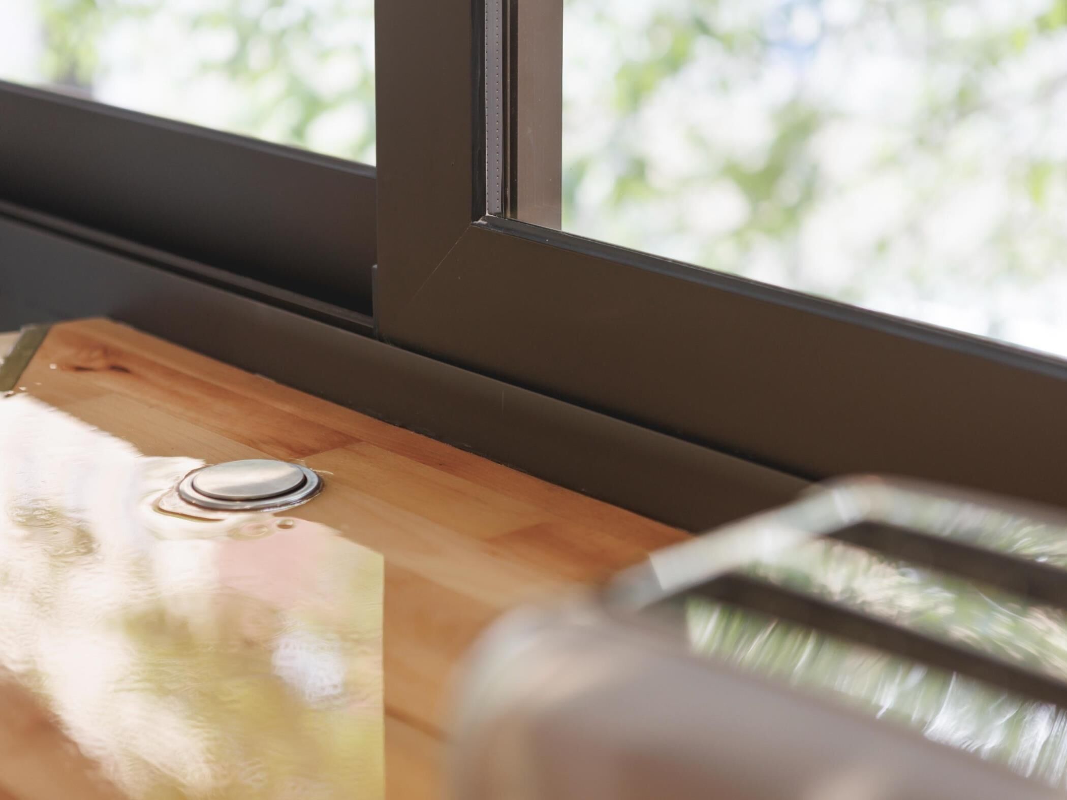 A wooden table with a metallic object, possibly a bottle, and a window with a view of greenery.