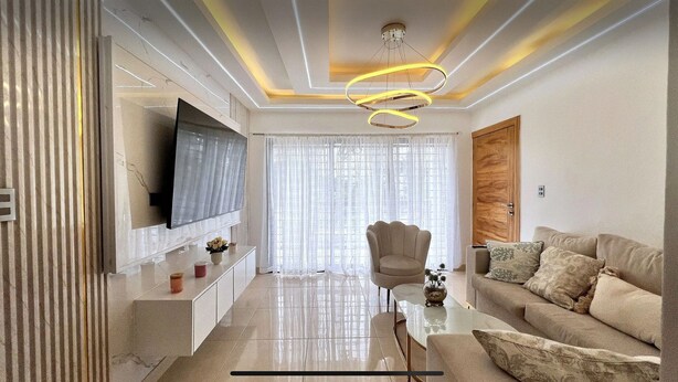 A modern living room with a flat-screen TV, a glass coffee table, a beige sofa, and a decorative chair.