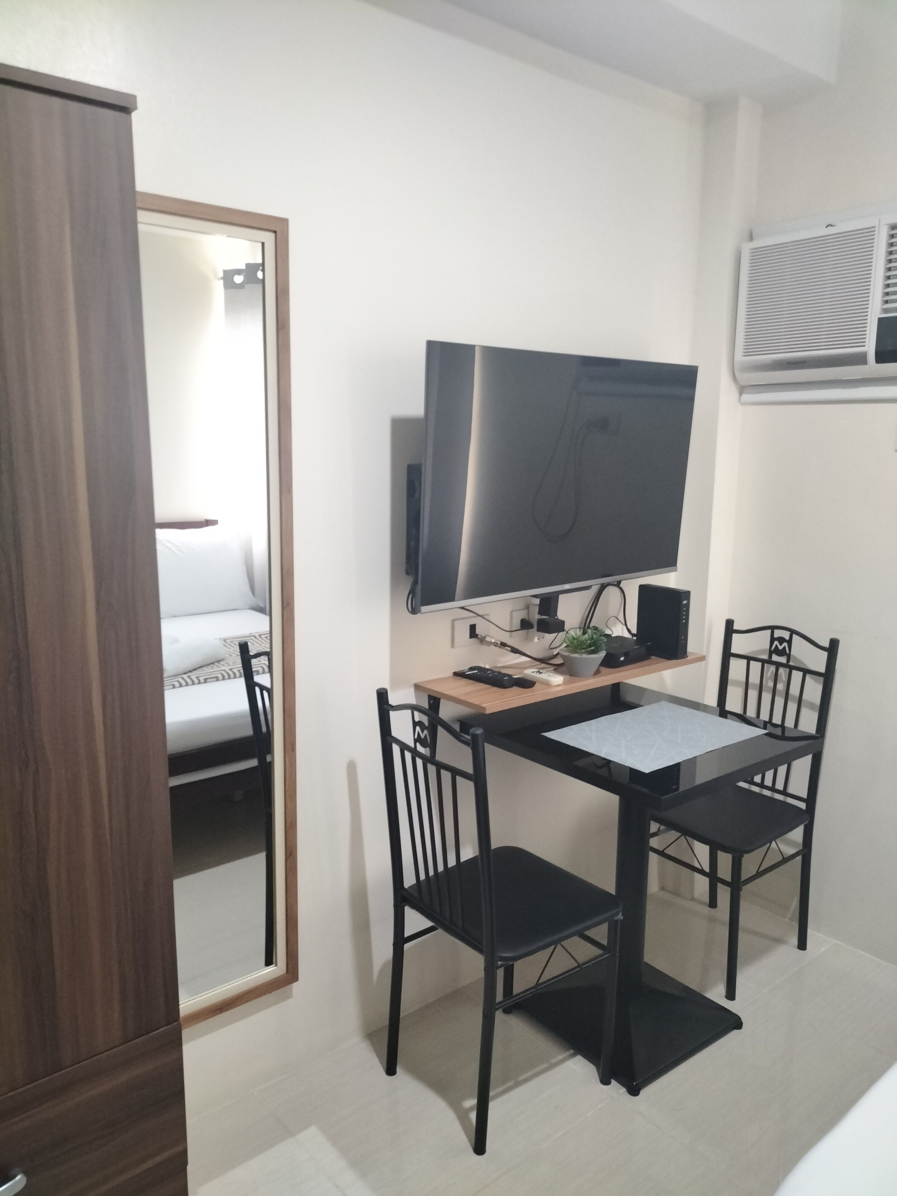A small, modern room with a wooden desk, two black chairs, a flat-screen TV, a mirror, and a bed with white bedding.