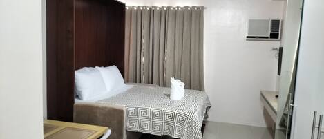 A hotel room with a bed, a desk, and a chair. There is a window with curtains, a wall-mounted air conditioner, and a mirror.