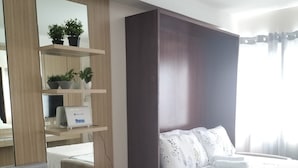 Een hotelkamer met een bed, een bureau met een computer, een stoel, een spiegel en planken met planten.