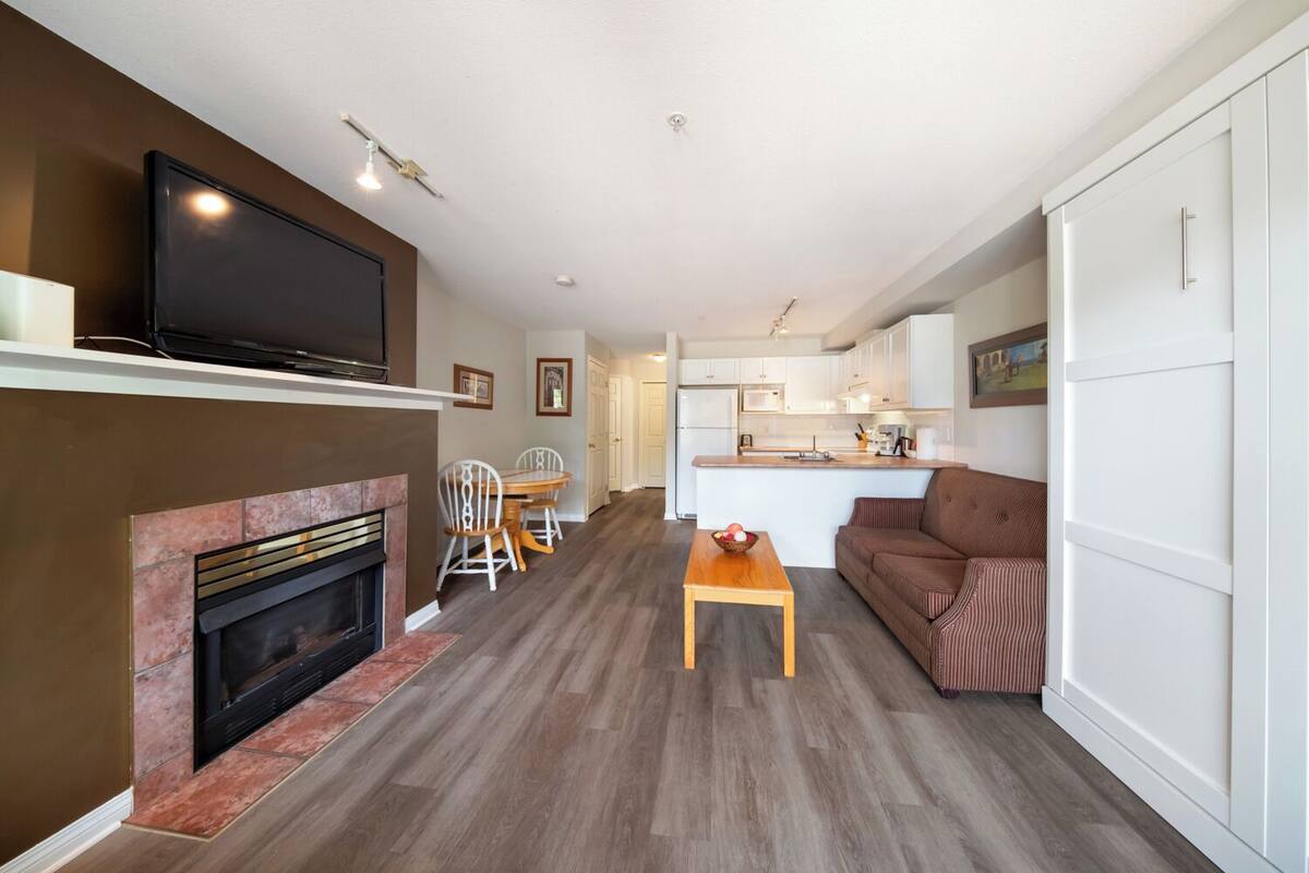 A modern living room with a fireplace, a television, a dining area, and a kitchen in the background.