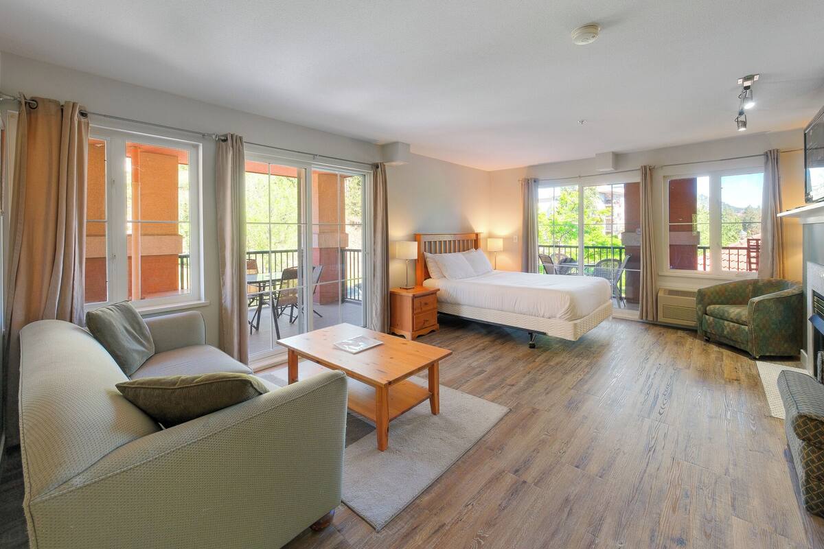 A hotel room with a sofa, a bed, a nightstand, a table, and a chair. There are large windows and a balcony.