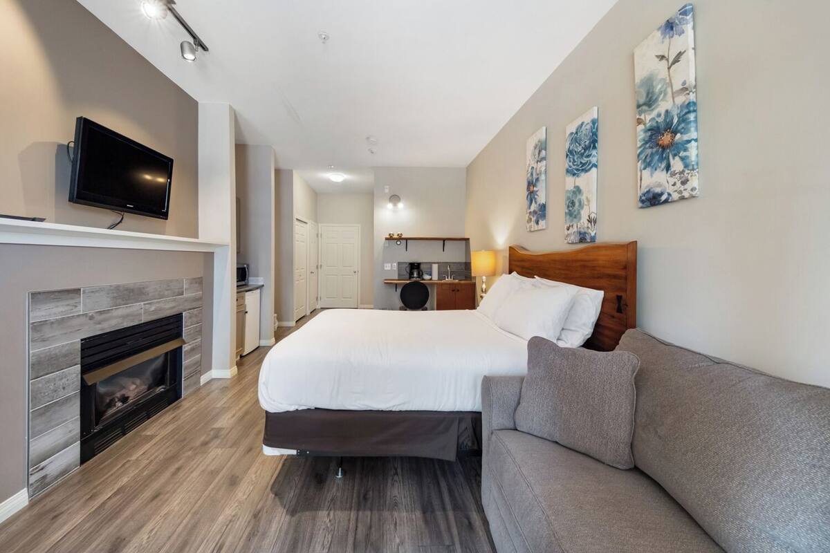 A modern hotel room with a large bed, a sofa, a fireplace, a television, and artwork on the walls.
