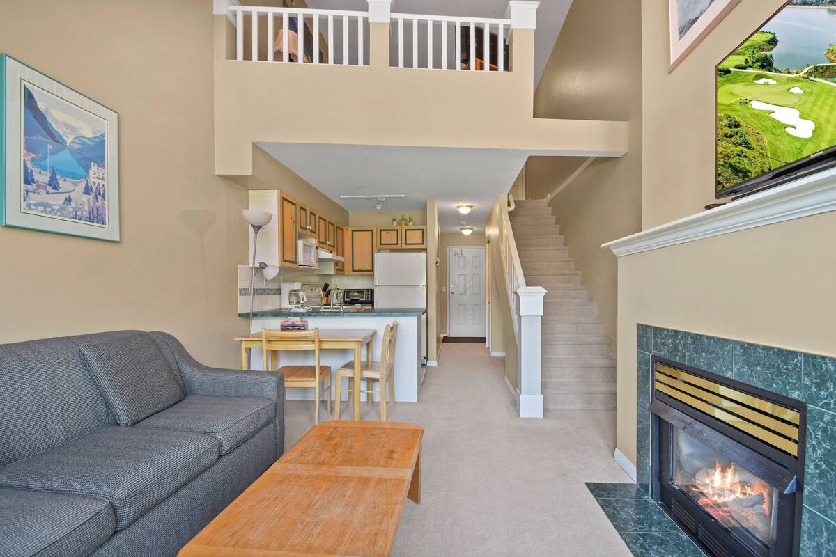 A living room with a sofa, coffee table, fireplace, and a view of the kitchen and stairs.