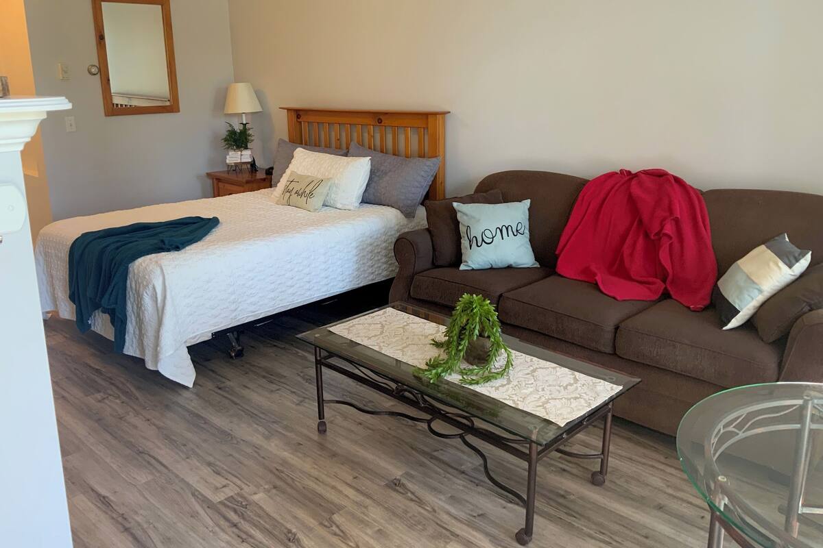 A bedroom with a bed, a nightstand, a dresser, a glass coffee table, and a brown sofa with a red blanket.