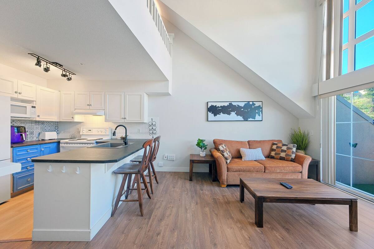 A modern kitchen with white cabinets, a central island, and a dining area with wooden chairs. There is a sofa and a coffee table in the living area, and a large window providing a view outside.