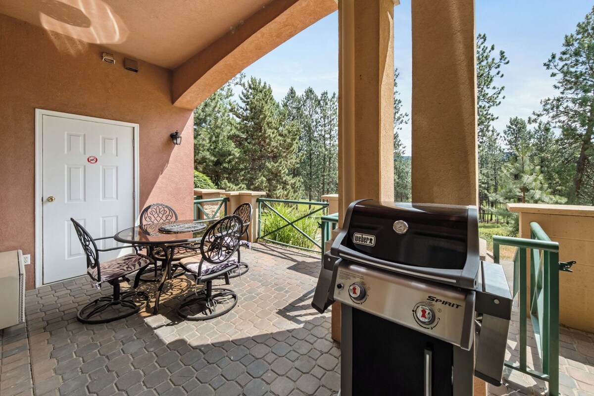 An outdoor patio with a Weber Spirit grill, a dining table with chairs, and a view of trees in the background.
