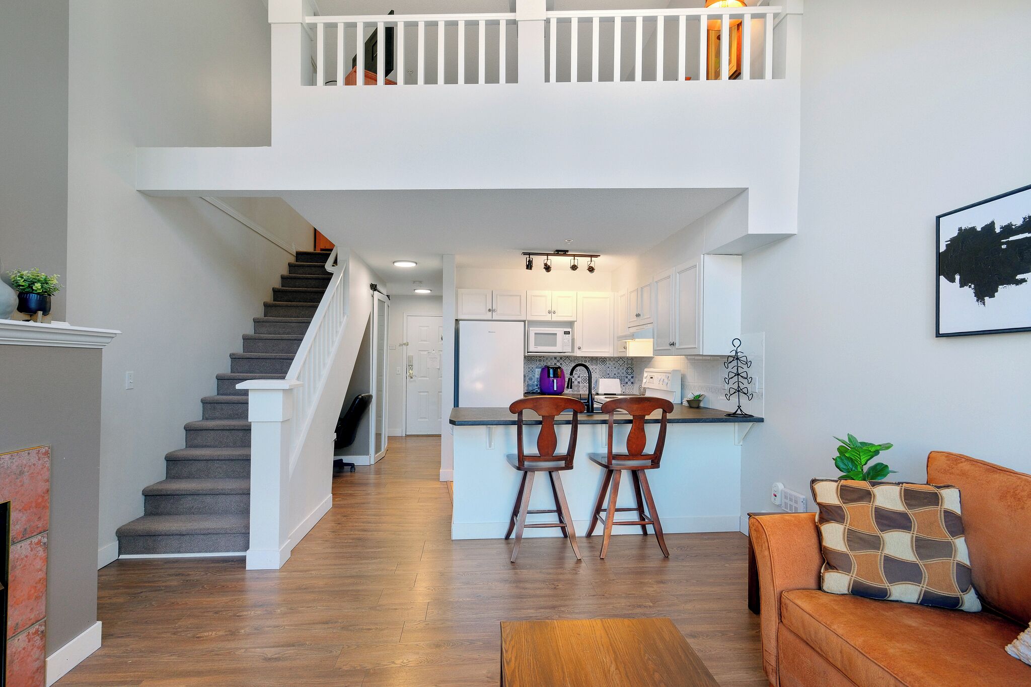 Un intérieur moderne avec un escalier menant à une mezzanine, une cuisine dotée de meubles blancs et un coin salon avec un canapé et des fauteuils.