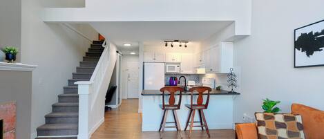 Un intérieur moderne avec un escalier menant à une mezzanine, une cuisine dotée de meubles blancs et un coin salon avec un canapé et des fauteuils.