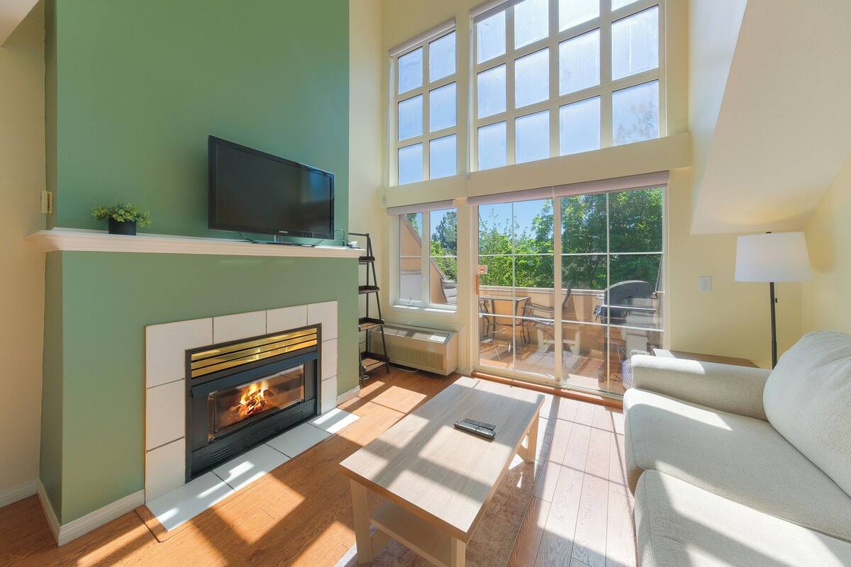 A living room with a fireplace, a television, a sofa, and large windows.