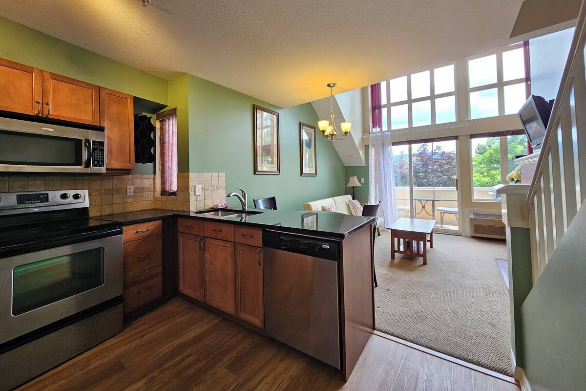 A kitchen with wooden cabinets, a microwave, an oven, and a dishwasher. There is a dining area with a table and chairs, and a large window providing a view of the outdoors.