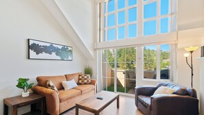 Ein Loft-Wohnzimmer mit hoher Decke, einem braunen Ledersofa, einem Holztisch und einem großen Fenster mit Blick ins Freie.