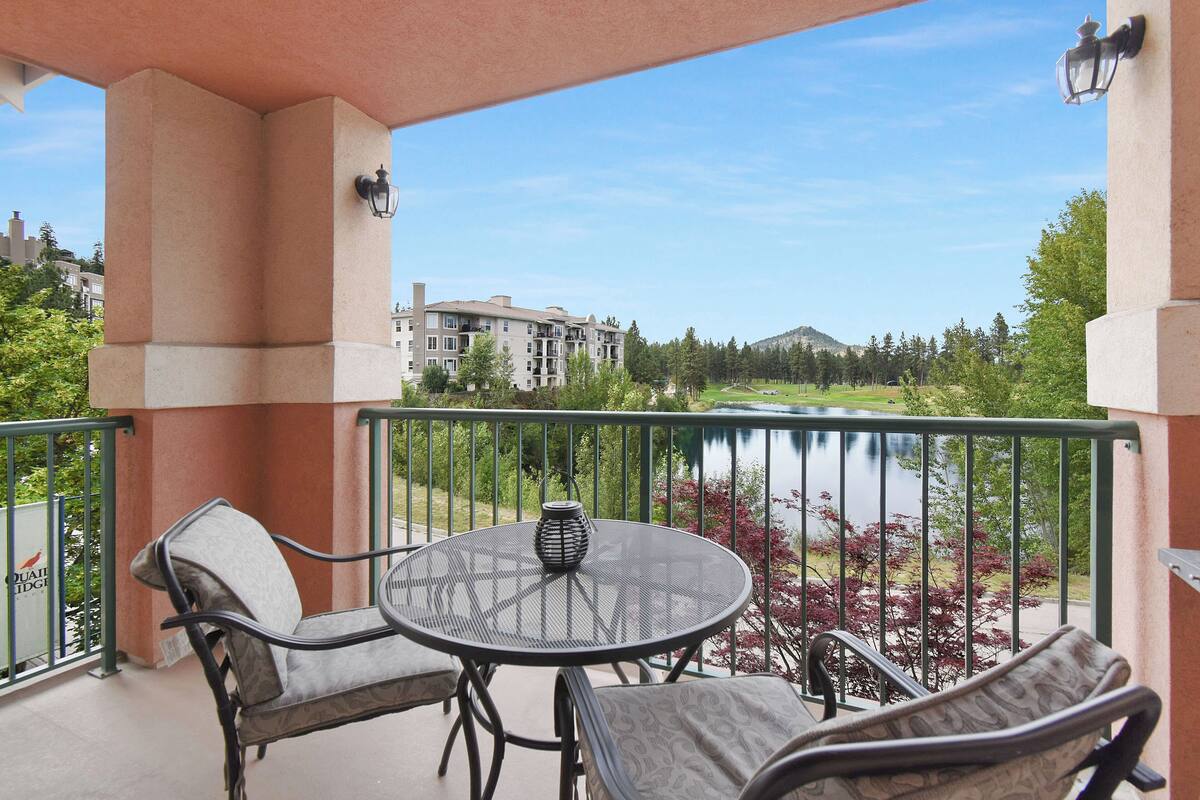 A balcony with a round table and two chairs, overlooking a lake and buildings.