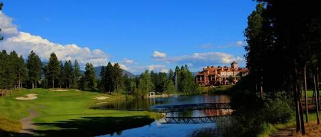 A golf course with a pond, a bridge, and a large building in the background.