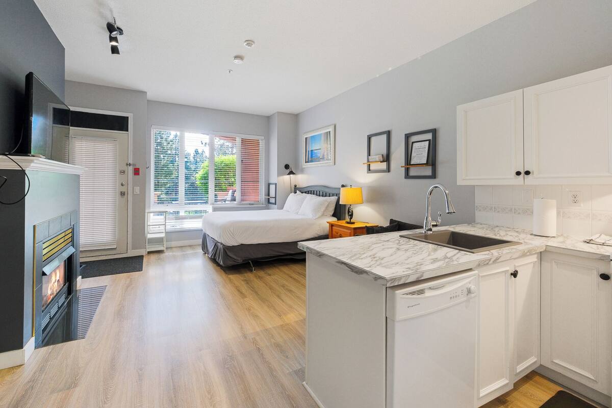 A modern hotel room with a bed, a small desk with a sink, a flat-screen TV, and a fireplace.