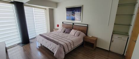 A bedroom with a large bed, a nightstand, a desk, a chair, a window with blinds, and a cityscape painting on the wall.